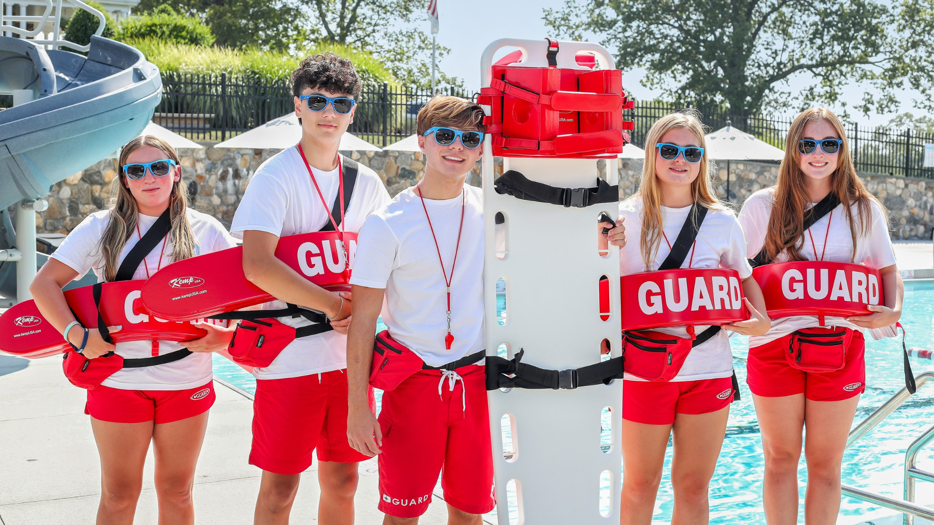 Group of five lifeguards smiling and posing with the stretcher by the pool
